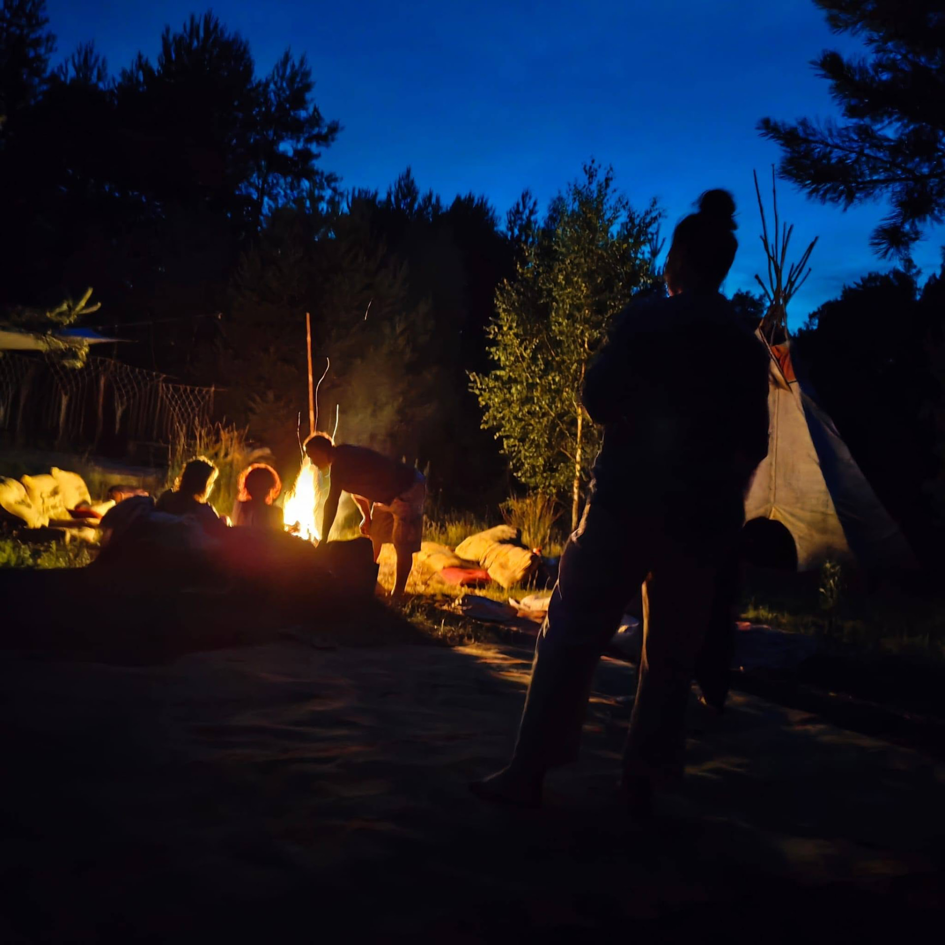 Ognisko o zmierzchu, sylwetki ludzi i tipi na polanie